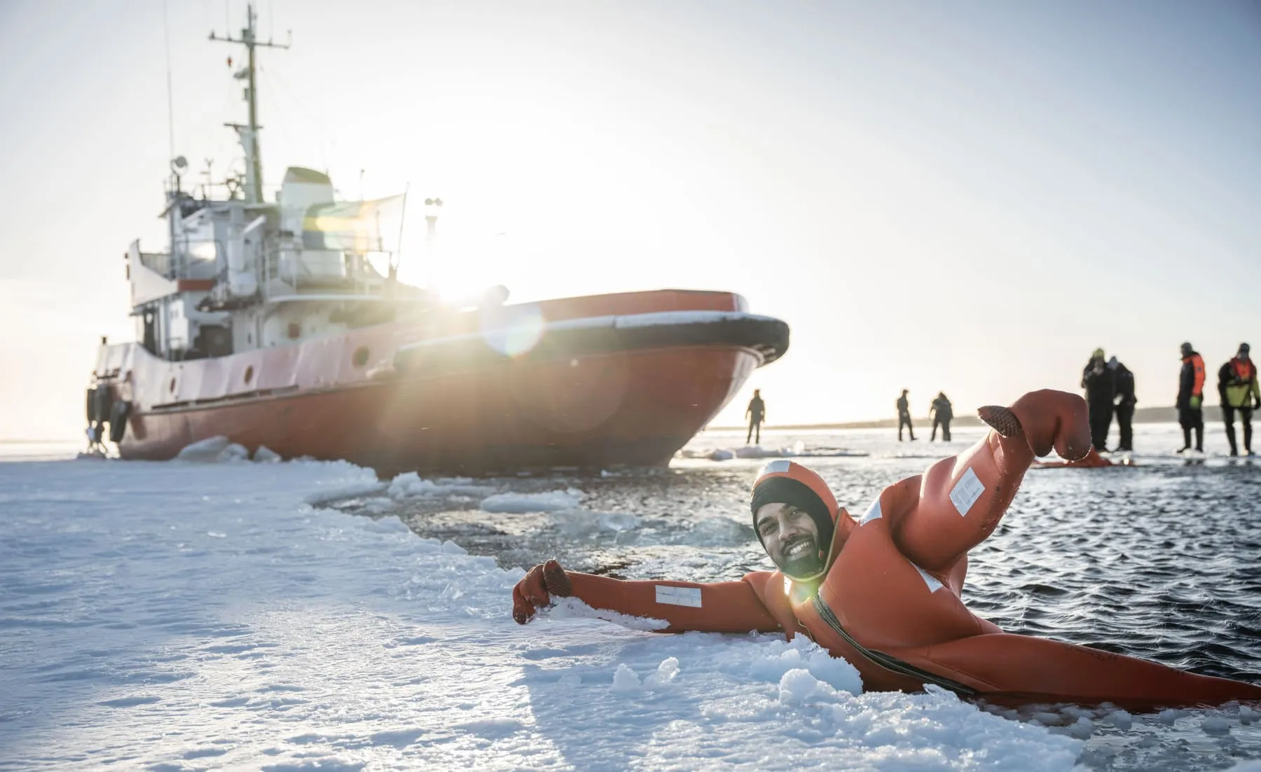 Man in orange survival suit lying on ice near open water with a large red icebreaker ship and group of people in the background.