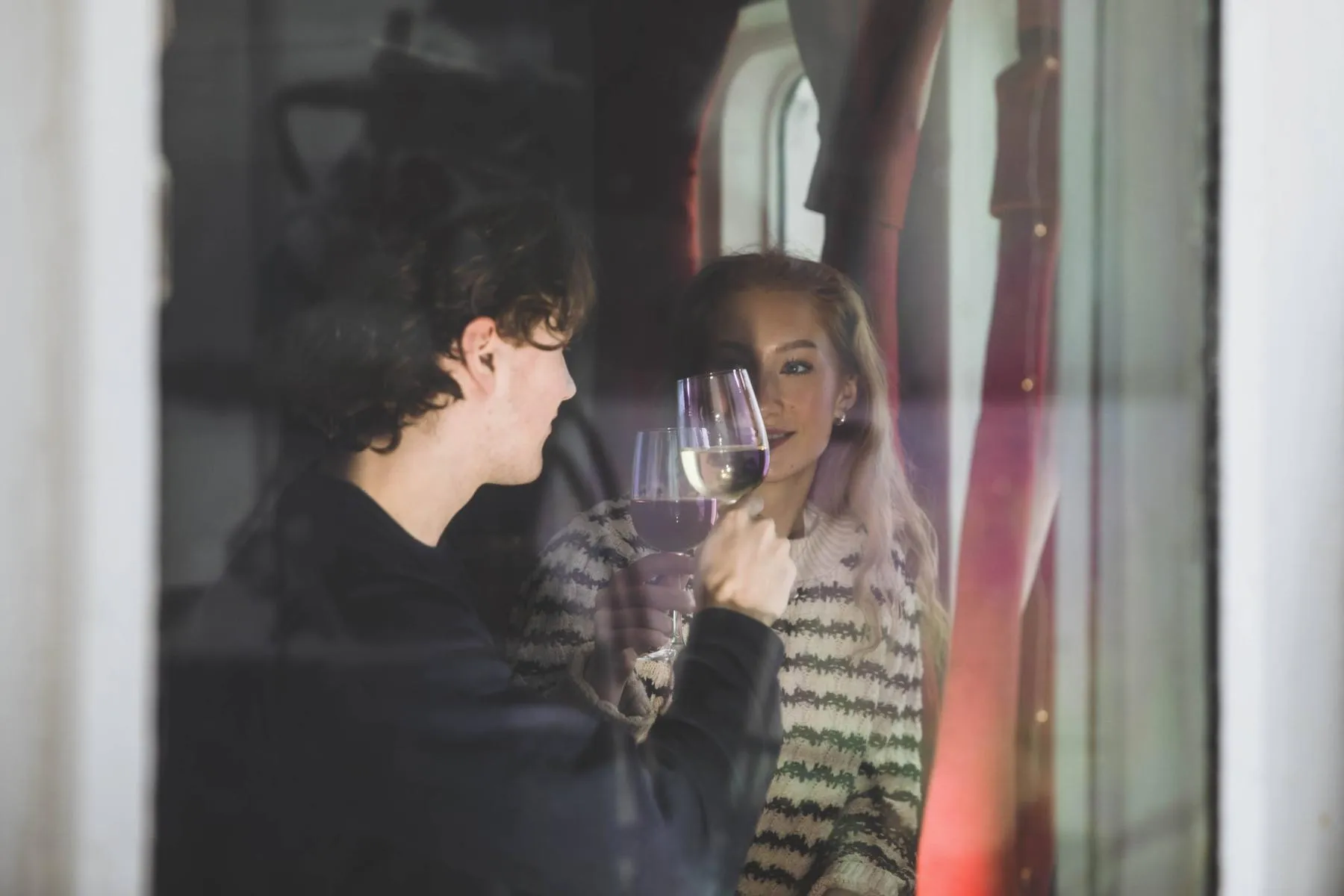 Man and woman clinking wine glasses inside, seen through a window.