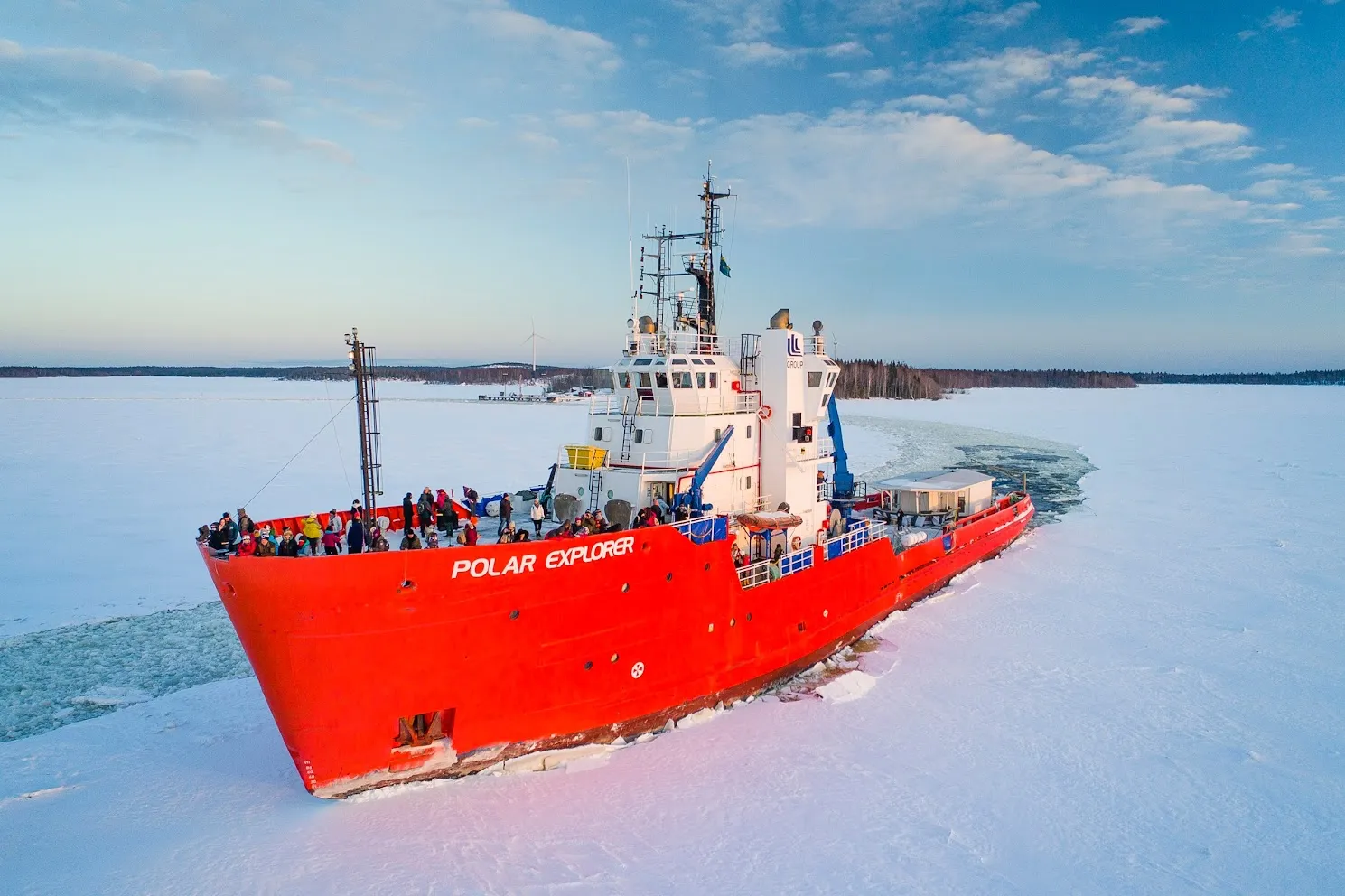 Red icebreaker ship named Polar Explorer breaking through frozen sea with people on deck against a blue sky.