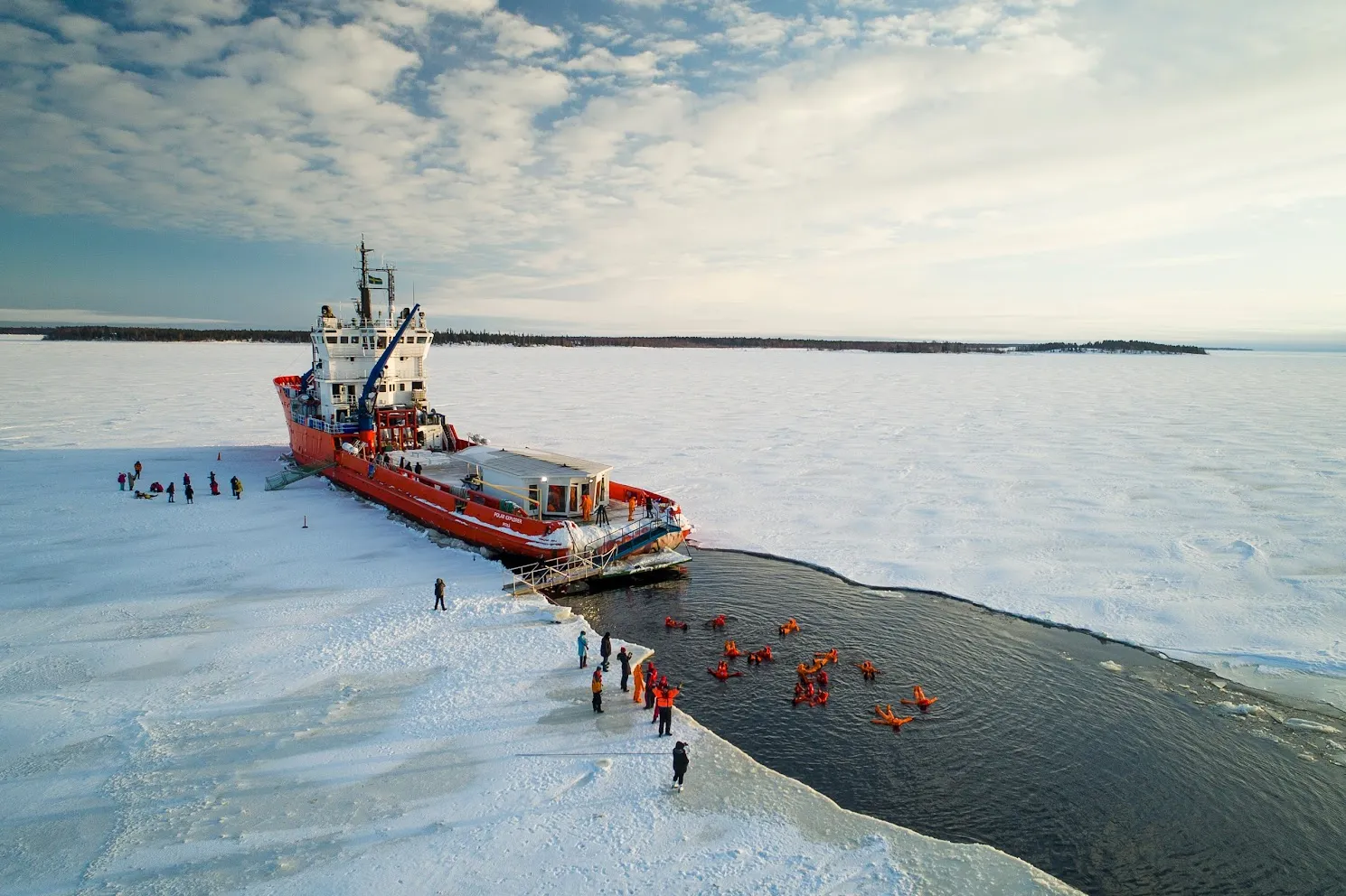 People in orange survival suits swimming in icy water next to a large red icebreaker ship on a frozen sea.
