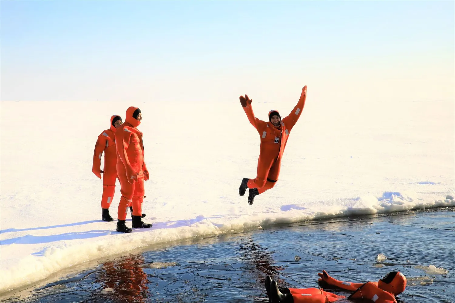 Four people in bright orange survival suits on ice near open water, with one person jumping into the water and another lying on the ice.