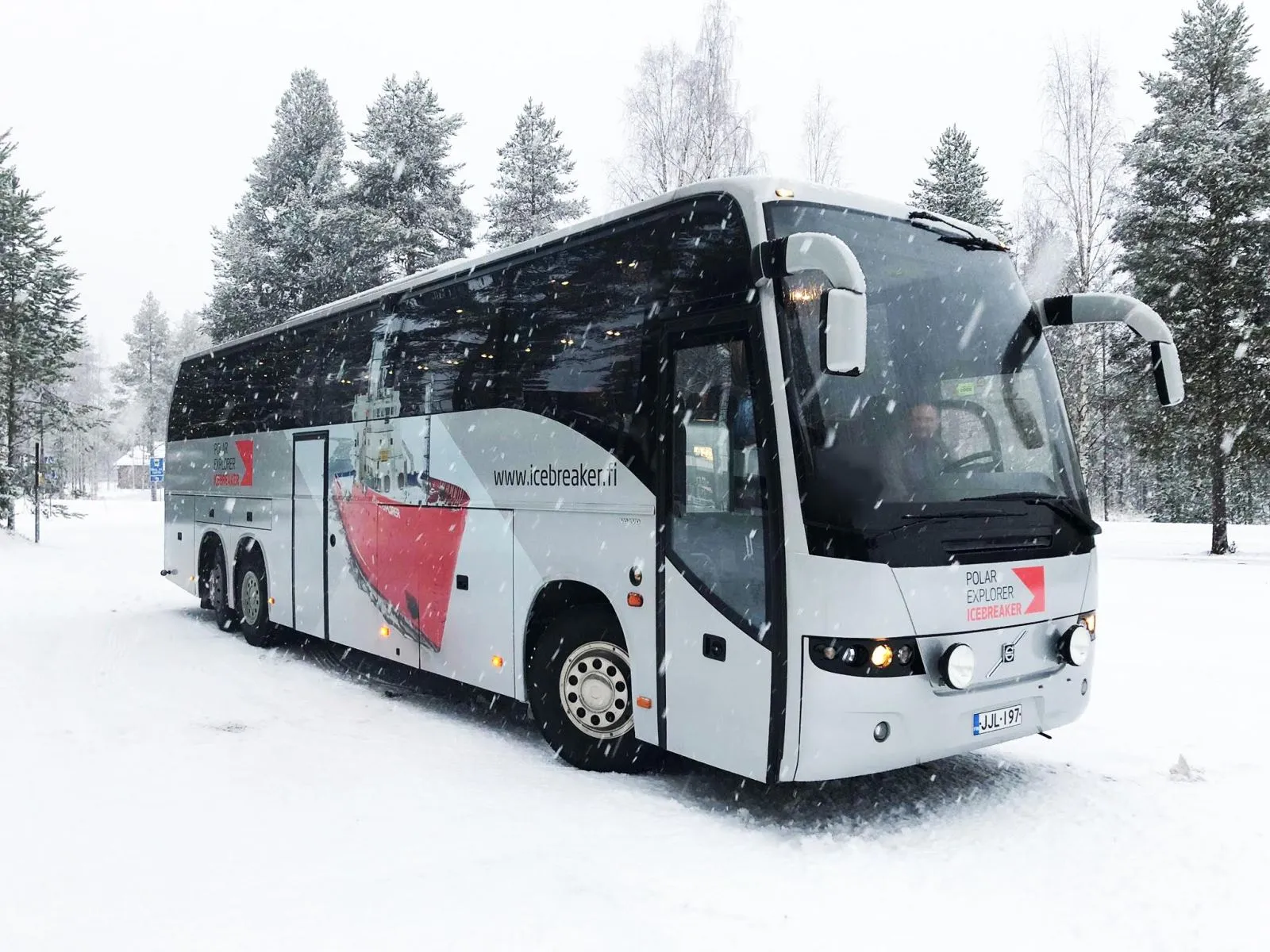 White and black shuttle bus with icebreaker ship image and text 'Polar Explorer Icebreaker' driving on snowy road surrounded by snow-covered trees.