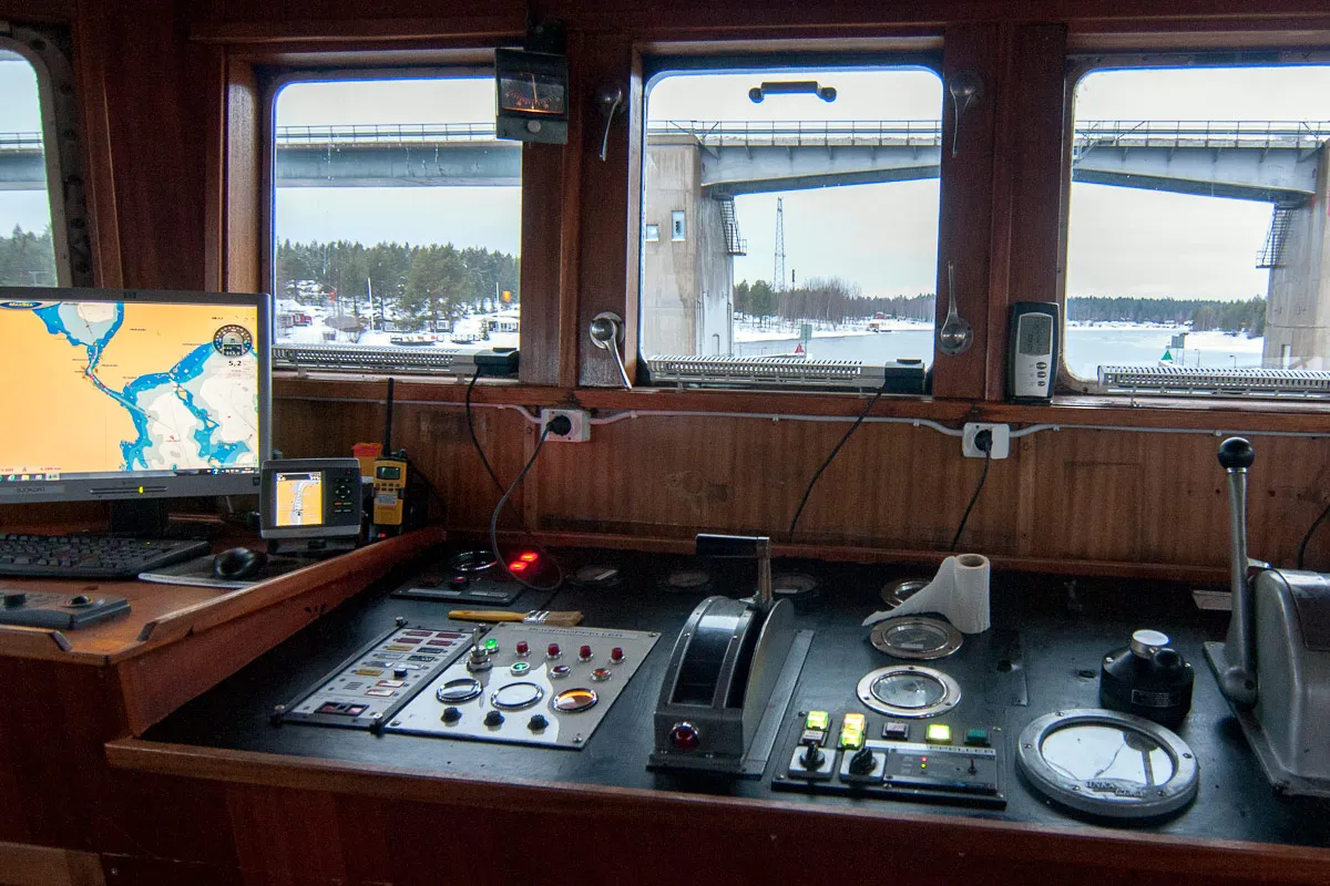 Control panel inside a ship's bridge with navigational instruments, computer screens showing maps, and windows overlooking a waterway with a bridge and snow-covered shore.