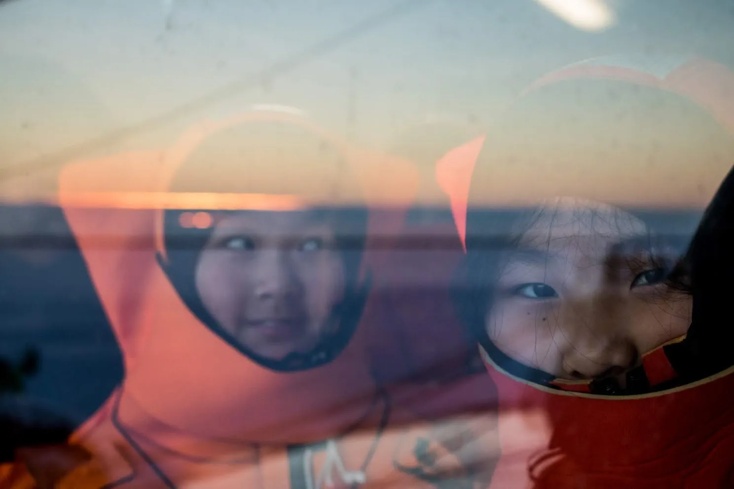 Two children wearing orange survival suits looking out through a rain-speckled window at sunset.