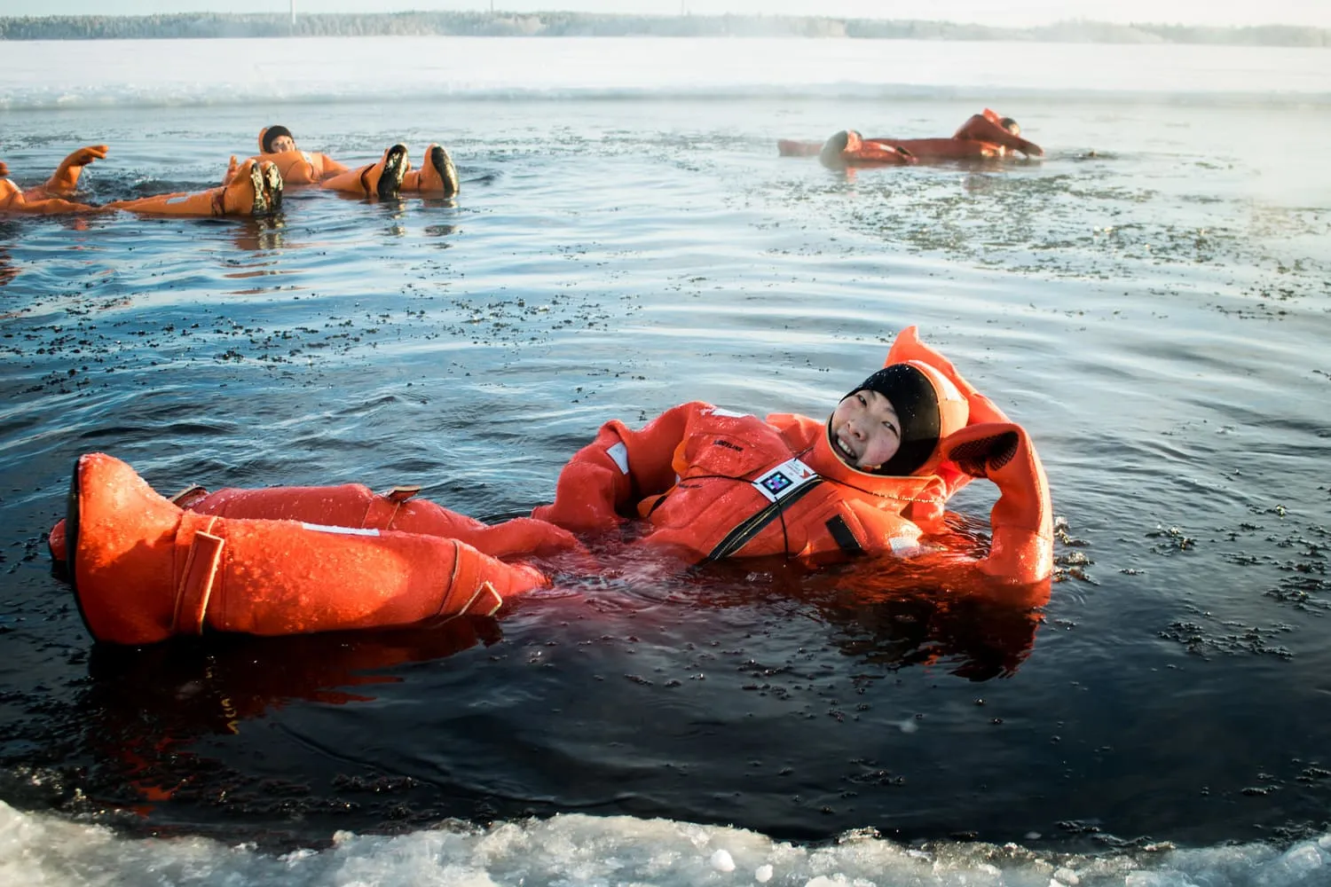 Person in an orange survival suit floating and smiling in icy water with other people in similar suits in the background.